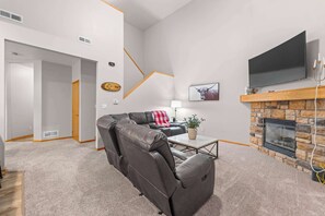 A modern living room with a leather sofa, a flat-screen TV mounted above a stone fireplace, and a hallway leading to another room.