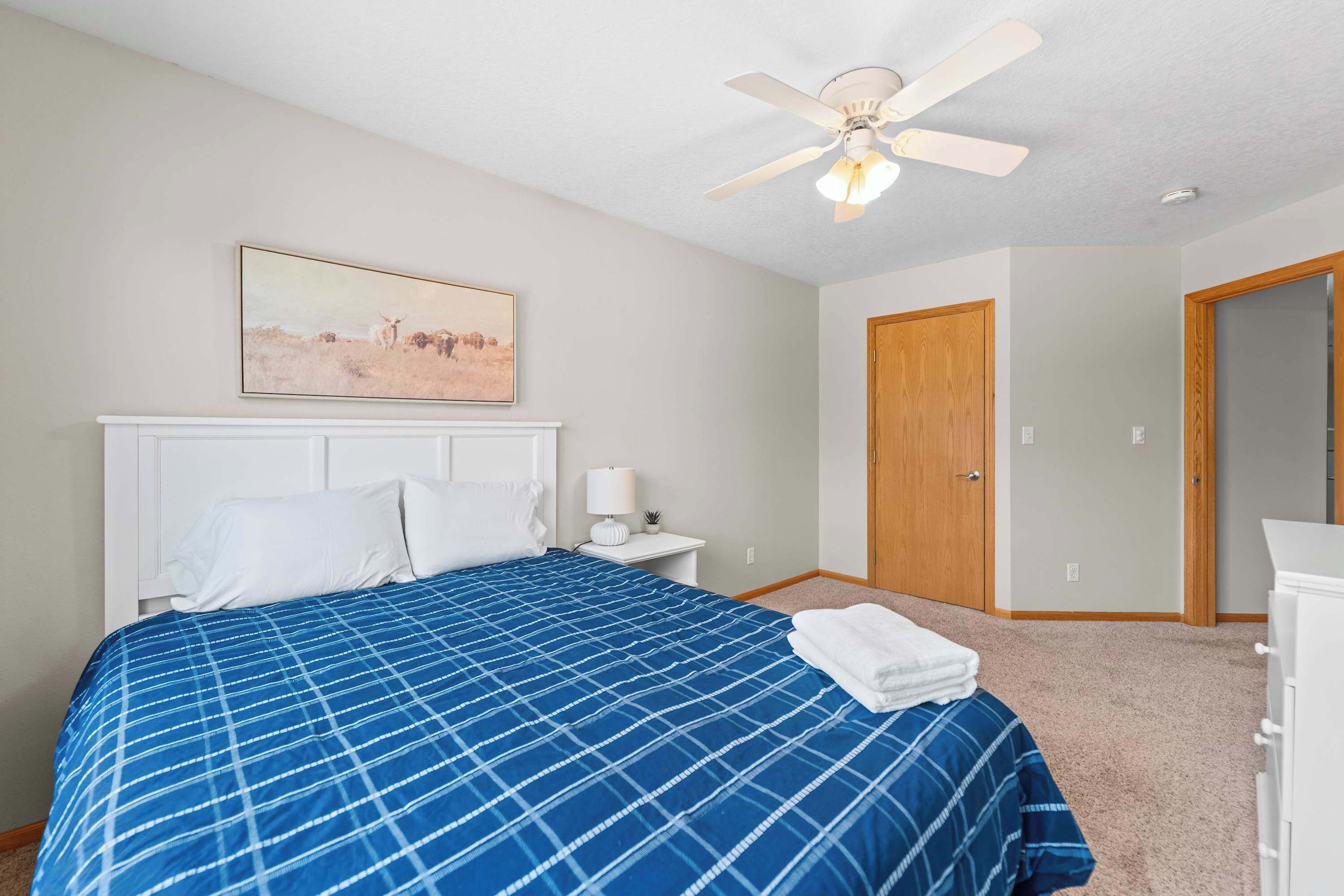 Une chambre à coucher comprenant un lit, un ventilateur de plafond, une table de chevet, une lampe, une porte et un tableau accroché au mur.
