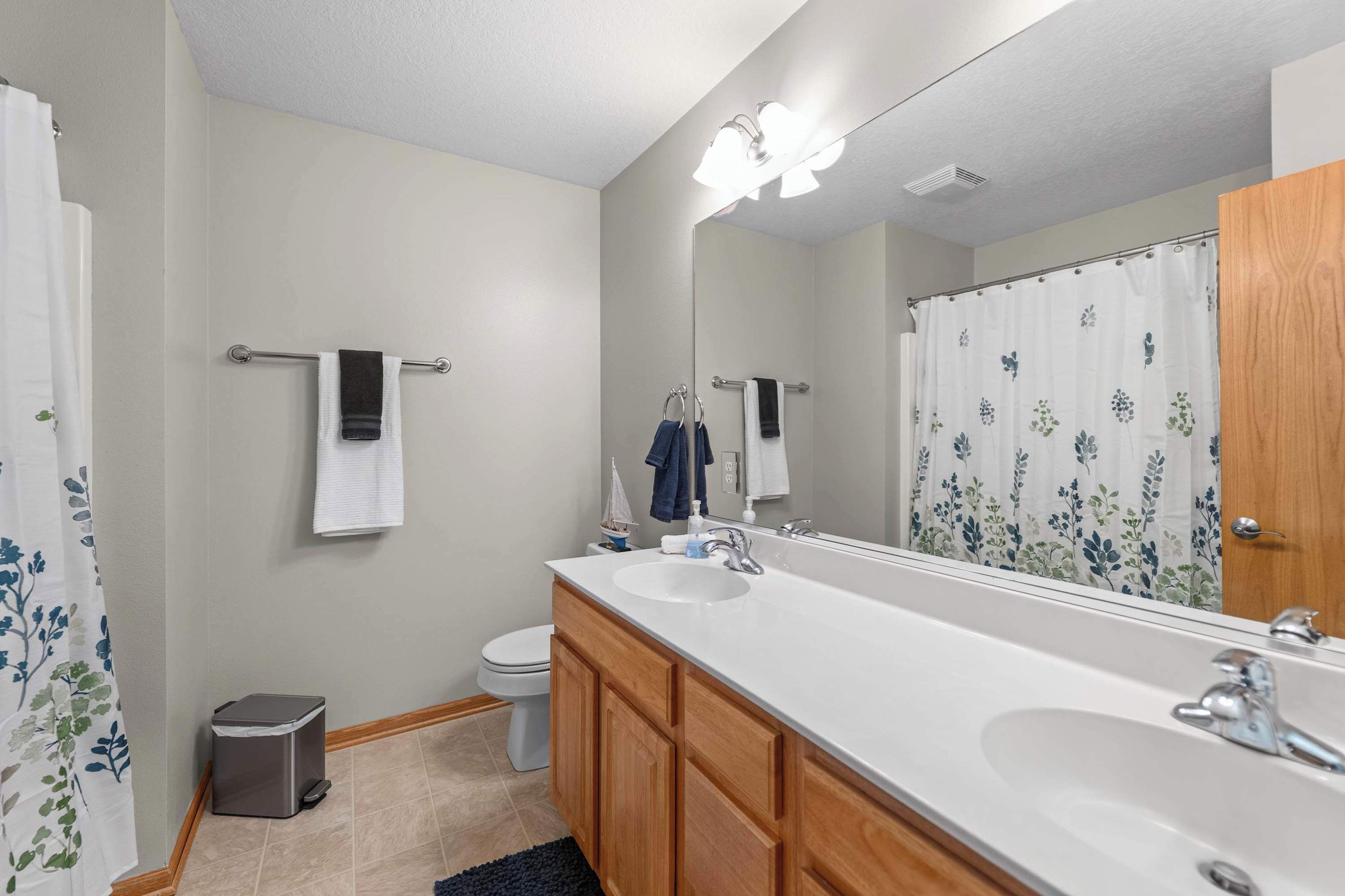 A bathroom with two sinks, a toilet, a shower curtain with a floral pattern, and a mirror with lights above.