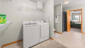 A laundry room with two white washers and dryers, a shelf with a detergent box, and a door leading to a bedroom.