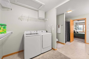 A laundry room with two white washers and dryers, a shelf with a detergent box, and a door leading to a bedroom.