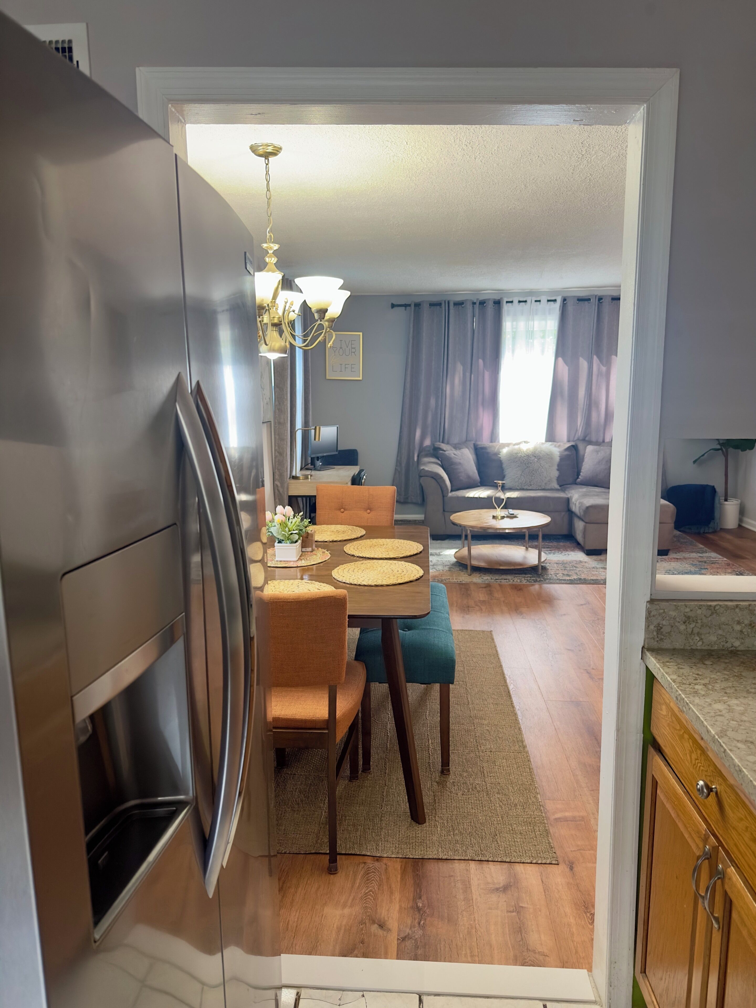 A modern kitchen with a dining area, wooden flooring, and a view into a living room with a sofa and curtains.