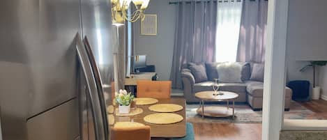 A modern kitchen with a dining area, wooden flooring, and a view into a living room with a sofa and curtains.