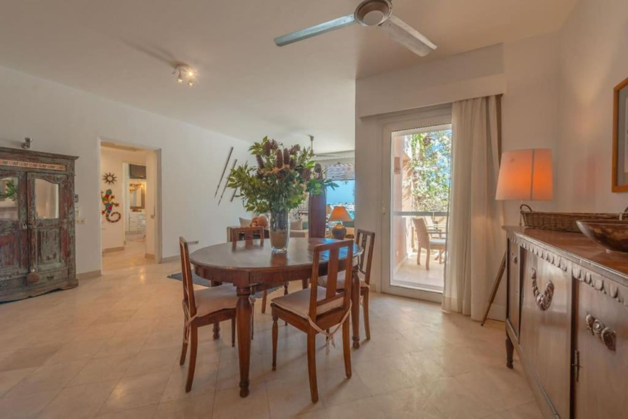 Un coin repas avec une table ronde et des chaises, un ventilateur de plafond et une vue sur une terrasse extérieure, accessible par une porte vitrée.