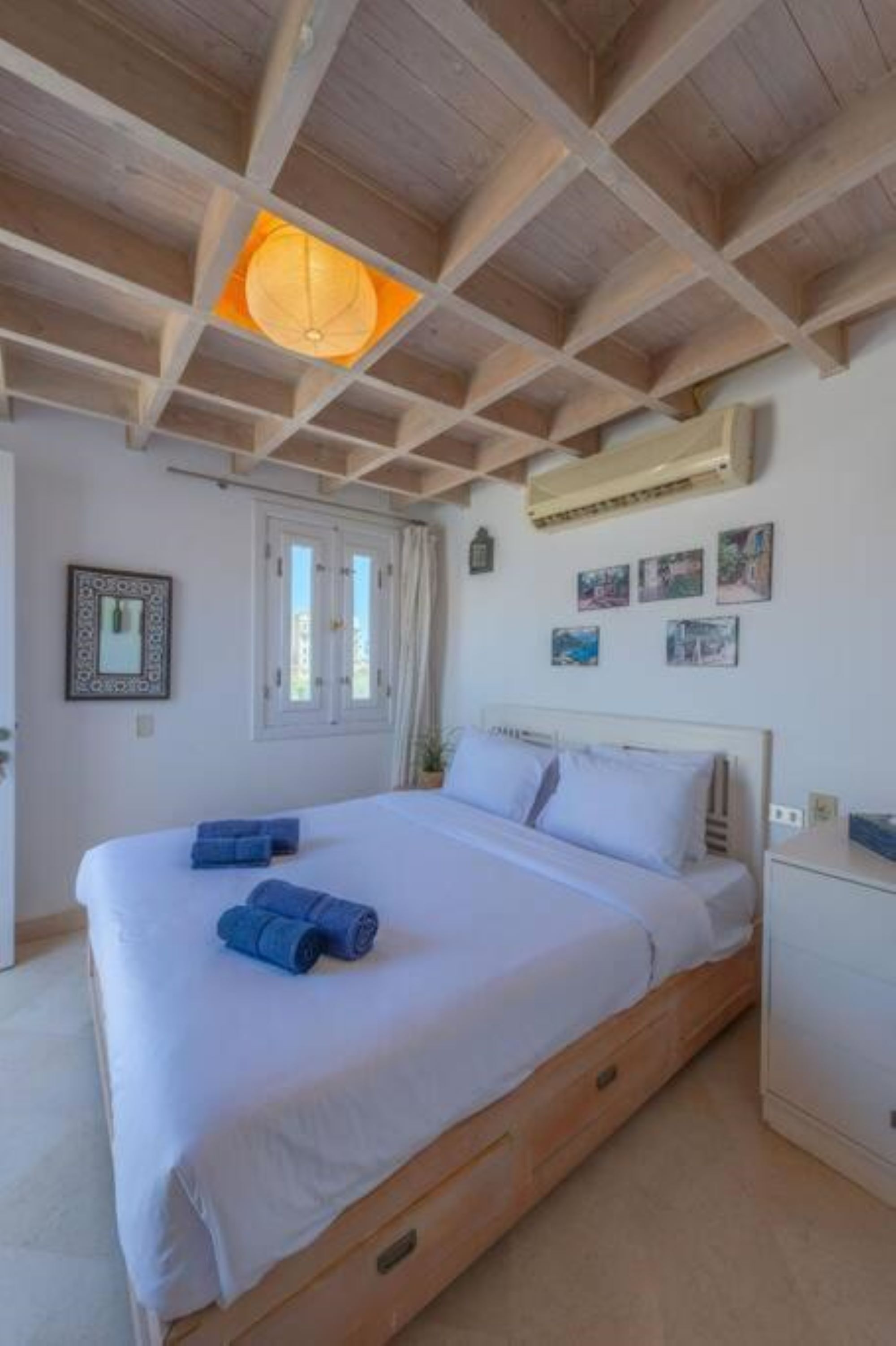 Une chambre avec un plafond en bois, un lit avec une literie blanche, une table de chevet et un climatiseur fixé au mur.
