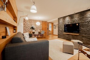 Een moderne woonkamer met een grijze bank, een eethoek met een tafel en stoelen, een flatscreen televisie bevestigd aan een stenen muur, en een houten plafond.