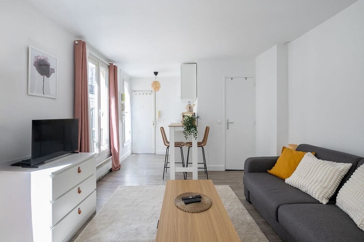 Un salon moderne avec un téléviseur à écran plat, un canapé gris agrémenté de coussins blancs et jaunes, une table basse en bois et un coin repas avec une table blanche et des chaises en osier.