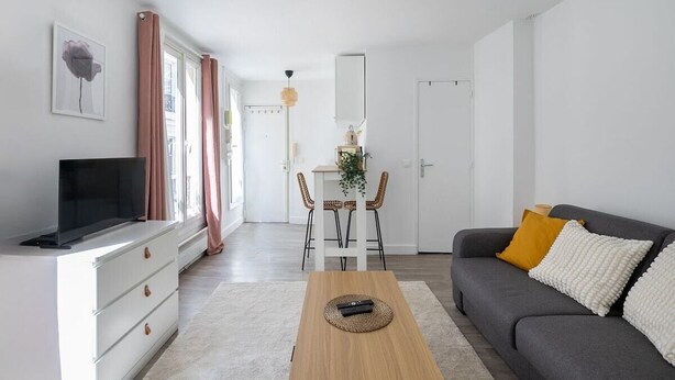 A modern living room with a flat-screen TV, a grey sofa with white and yellow cushions, a wooden coffee table, and a dining area with a white table and wicker chairs. - Studio in Pigalle - Moulin Rouge and Montmartre (Paris)