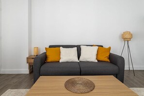 Un saló modern amb un sofà gris, dos coixins groc mostassa, una taula rodona de vimetes i una làmpada de peu.