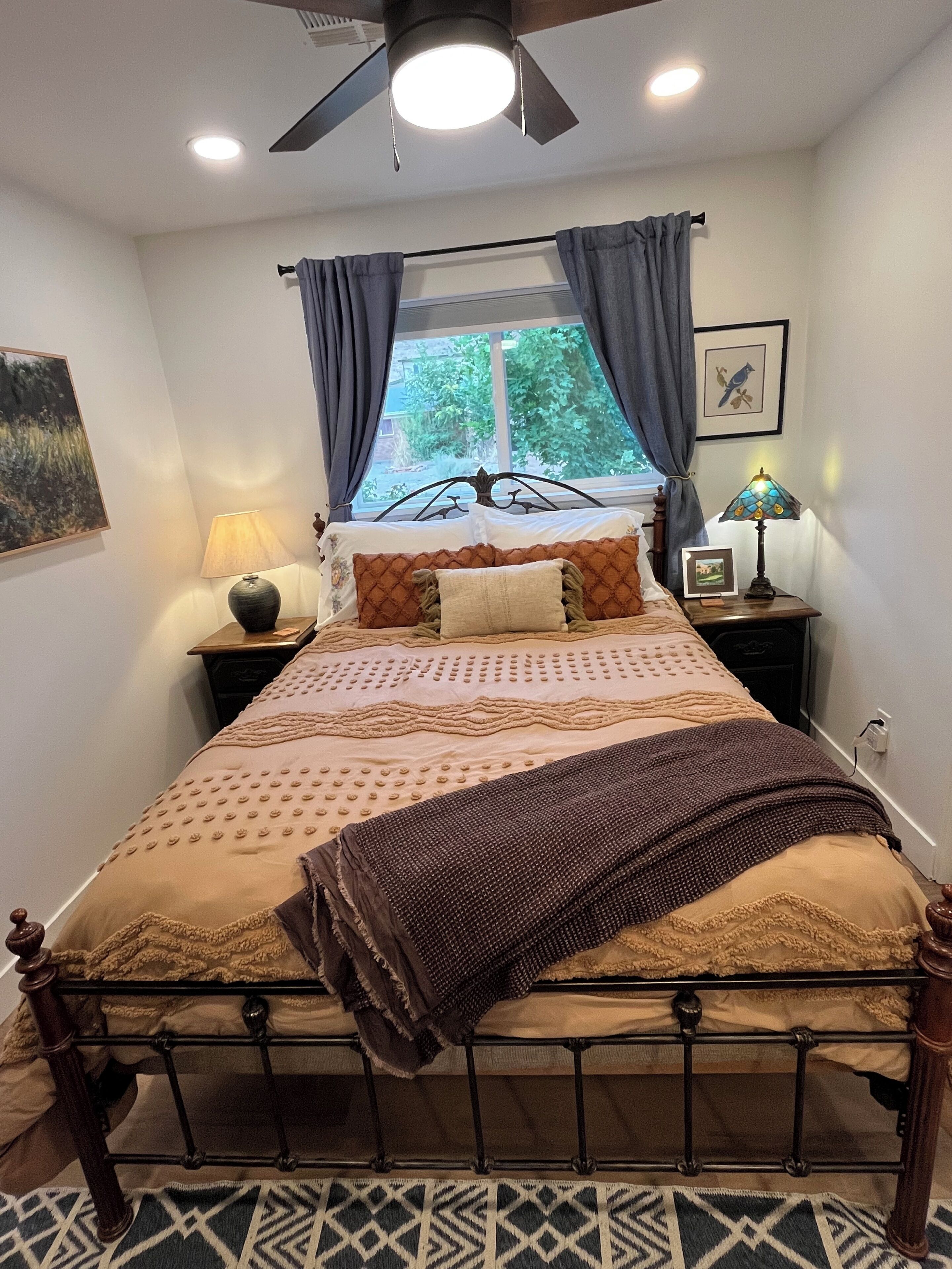 Un dormitorio con cama, ventana con cortinas, mesita de noche con lámpara y ventilador de techo.