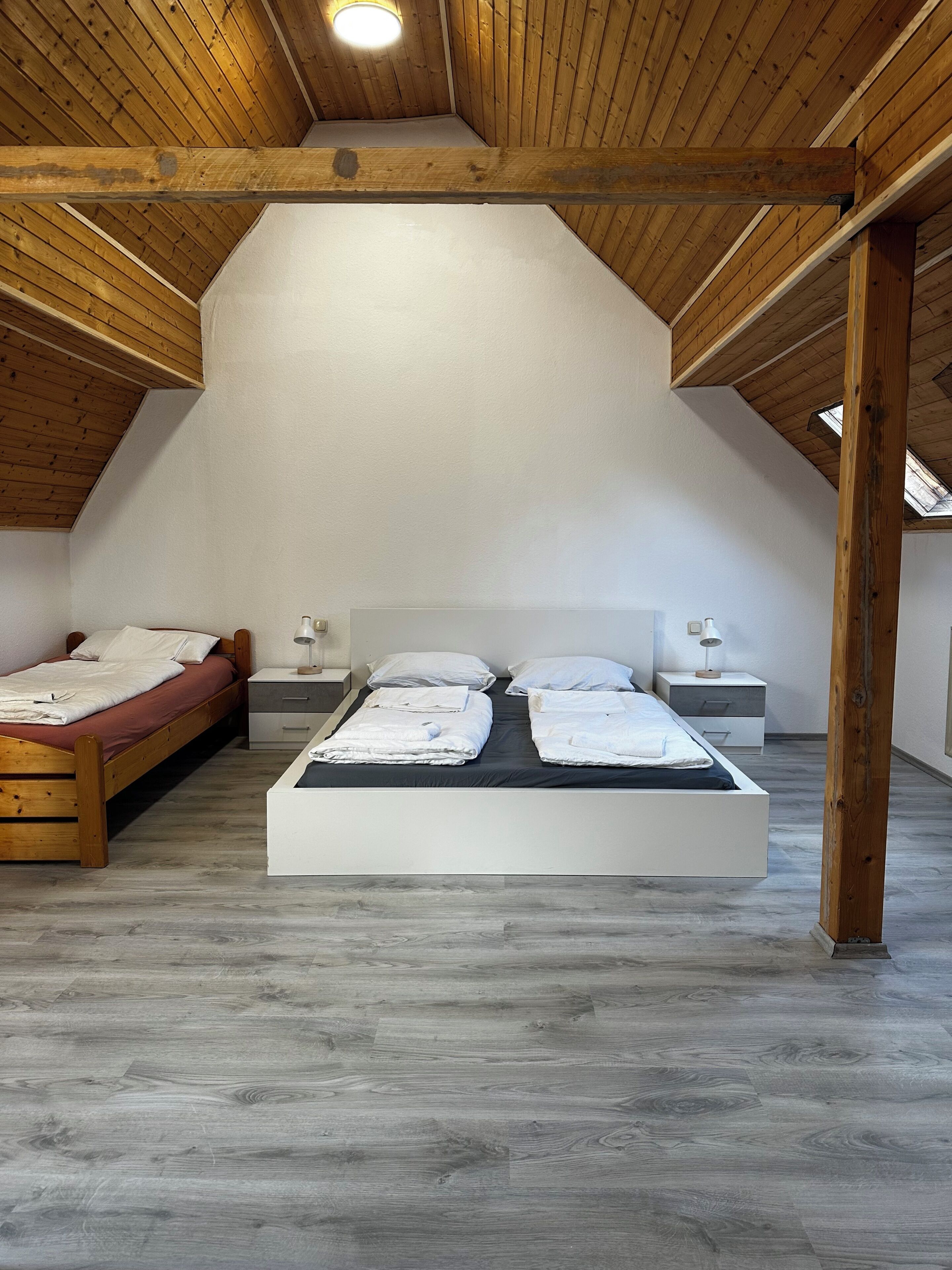 Une chambre avec un plafond en pente, des poutres en bois, un grand lit, des tables de chevet et une lucarne.