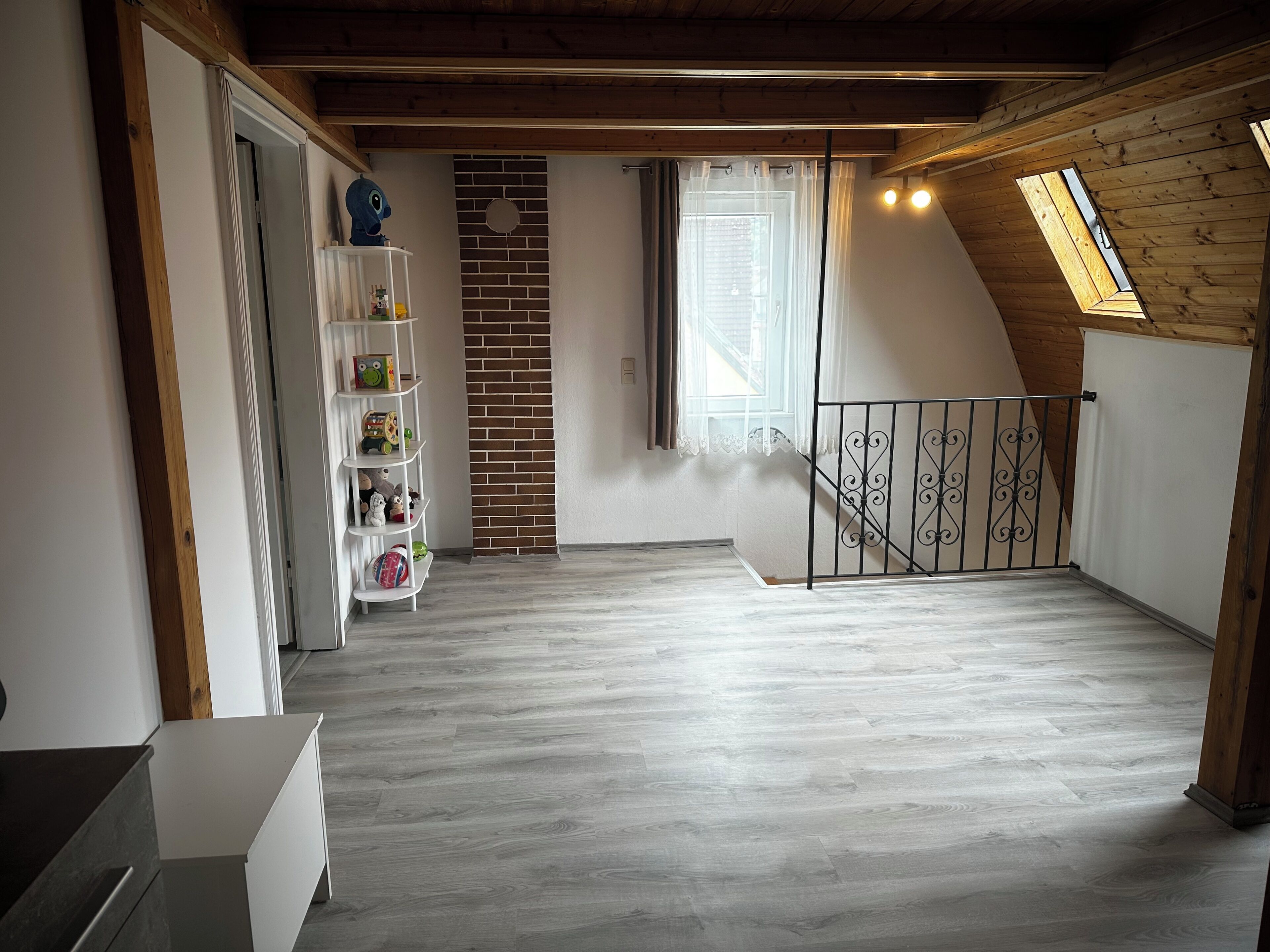 Une pièce avec un plafond en pente, un mur en briques, une étagère blanche remplie de jouets, une fenêtre avec des rideaux et un plancher en bois.