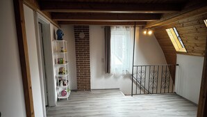 A room with a sloped ceiling, a brick wall, a white shelf with toys, a window with curtains, and a wooden floor.