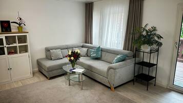 Ein Wohnzimmer mit grauem Sofa, einem Glastisch, einem Bücherregal und einem Blumentopf.
