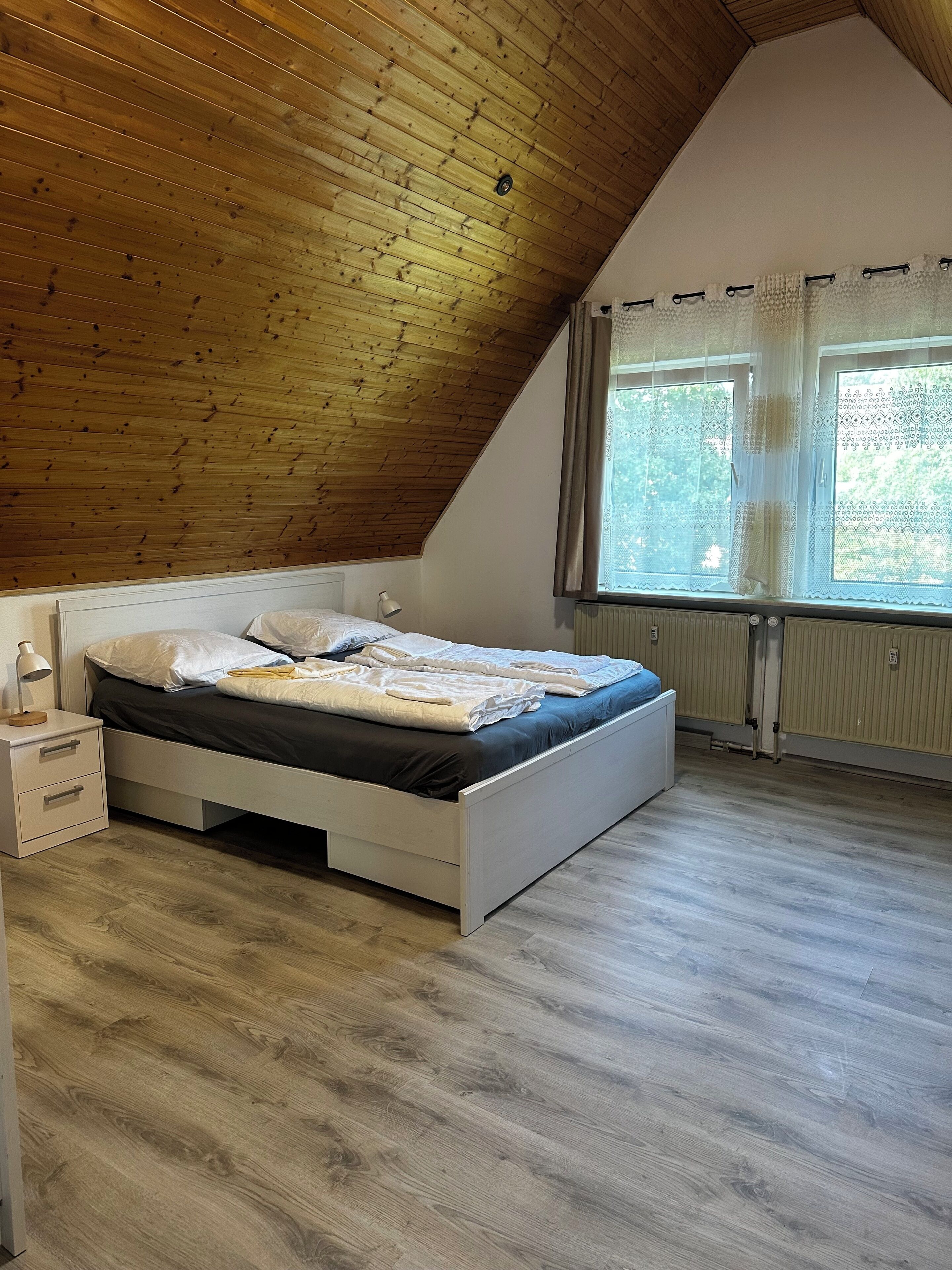 Ein Schlafzimmer mit schrägem Dach, einem Bett mit weißem Gestell, einem Nachttisch mit Lampe und einem Fenster mit Vorhängen.