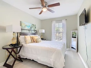 A bedroom with a bed, a ceiling fan, a nightstand, a lamp, a painting, a window with curtains, and a TV mounted on the wall. - Reunion Resort Getaway Close to Disney World. (Kissimmee)