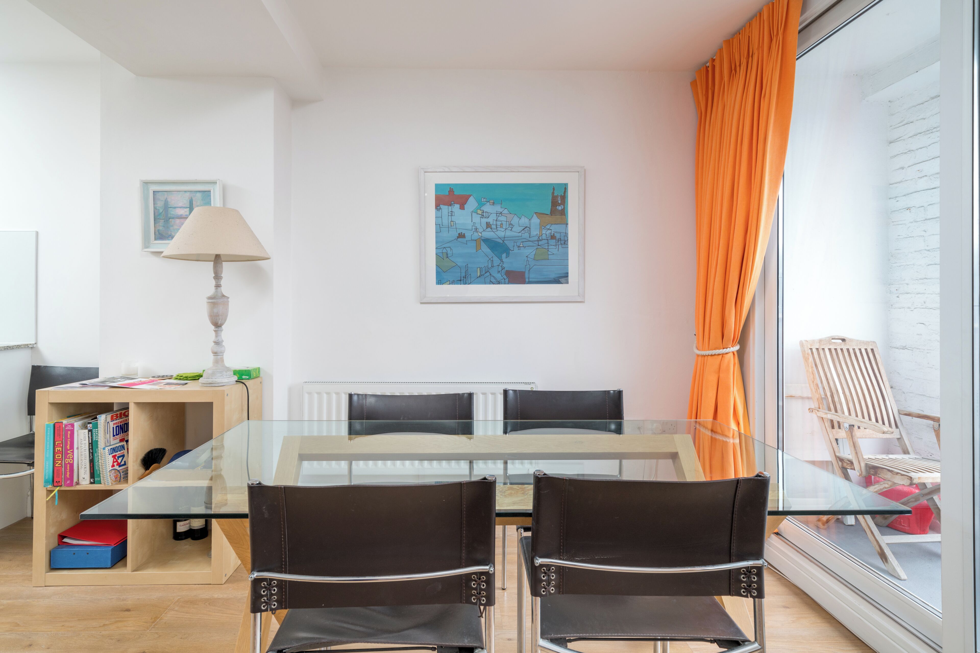 A modern living room with a glass dining table, two chairs, a bookshelf, a lamp, and a painting on the wall.