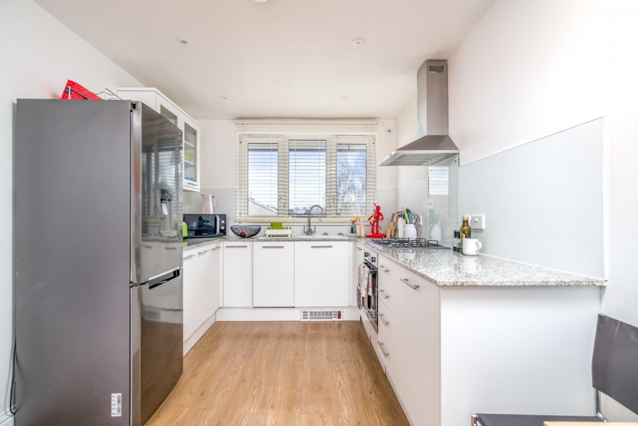 A modern kitchen with white cabinets, a stainless steel refrigerator, a built-in oven, and a window with blinds.