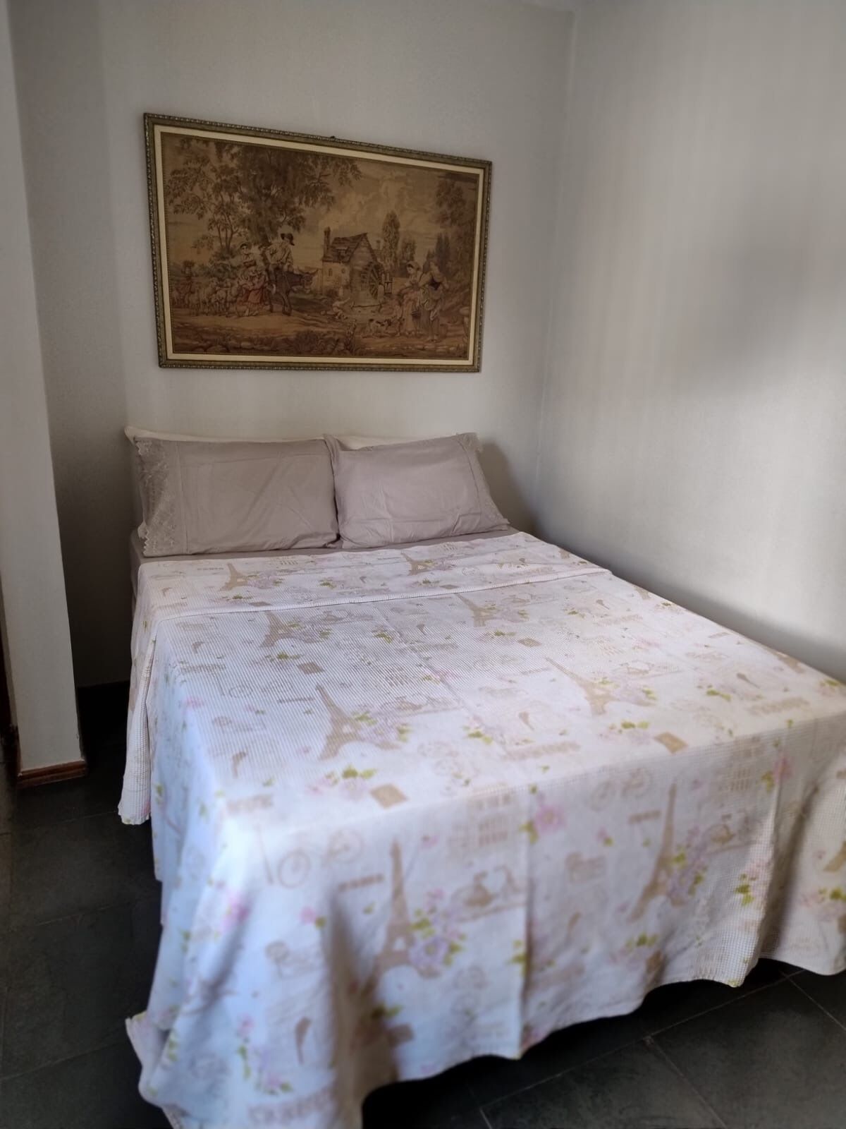A single bed with a floral bedspread, two pillows, and a framed landscape painting on the wall.