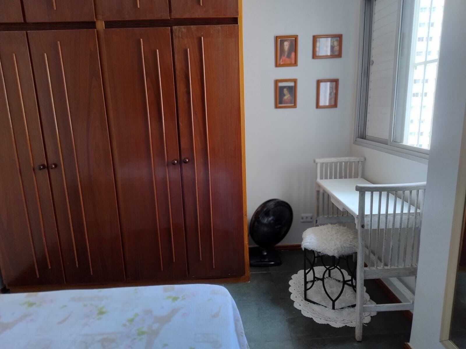 A bedroom with a wooden wardrobe, a crib, and a window with a view of buildings.