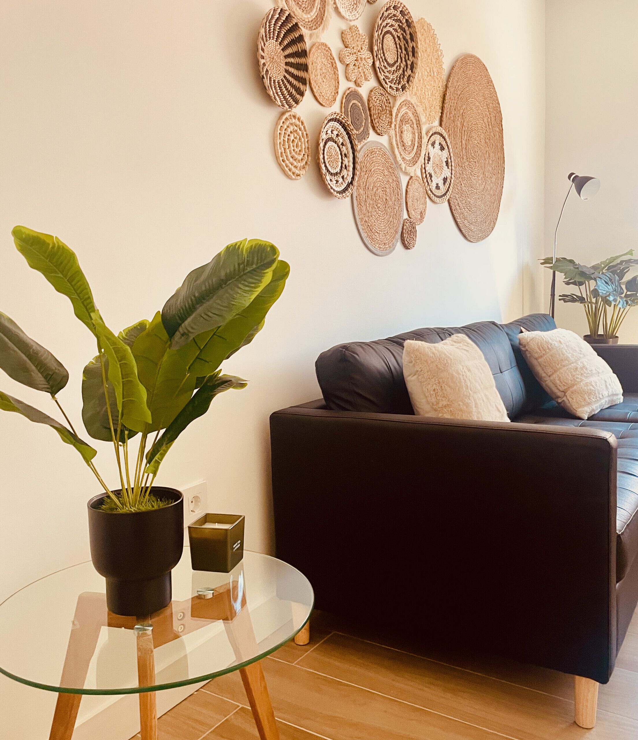 A modern living room with a glass coffee table, a dark sofa with cushions, a potted plant, and a wall art piece.