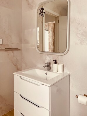 Un baño con tocador blanco, espejo, grifo, dispensador de jabón y porta papel higiénico.