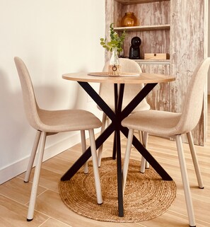 Una mesa redonda de madera con un mantel tejido, rodeada por cuatro sillas beige con patas negras, sobre un piso de madera.