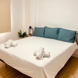 A neatly made bed with white linens and teal pillows, a small bedside table with a lamp, and a potted plant. - Click&Guest - Doctoral Urbana 1 (Santa Lucía de Tirajana)
