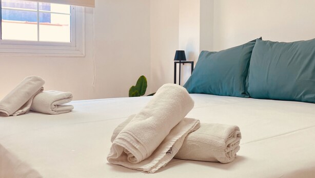 Ein ordentlich bezogenes Bett mit weißen Laken und aufgerollten Handtüchern, ein Fenster mit Jalousien, ein Blumentopf und blaue Kissen.