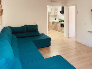 Ein smaragdgrünes Ecksofa in einem modernen Wohnzimmer mit Holzboden, einer Küche im Hintergrund und einer weißen Tür.
