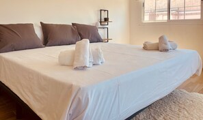 Ein ordentlich bezogenes Bett mit weißen Laken und braunen Kissen, ein gefaltetes Handtuch auf dem Bett, und ein Fenster mit Blick auf ein Gebäude.