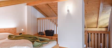Un dormitorio con techo de madera, cama con ropa blanca, mesita de noche y una lámpara fijada a la pared.