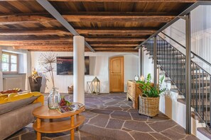 Un interior acogedor con suelo de piedra, vigas de madera, una escalera y una zona de estar con mesa y cojines.