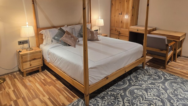 Una cama con dosel sobre un armazón de madera, una mesita de noche con lámpara, un guardarropa de madera y un escritorio con silla.
