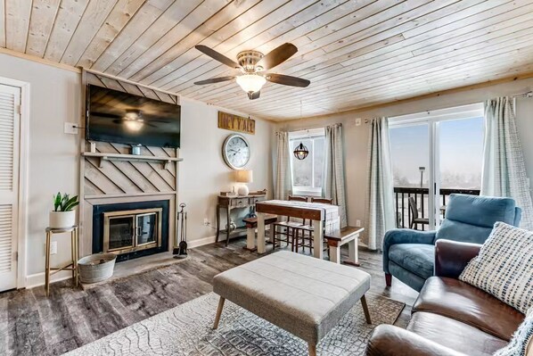 A cozy living room with a fireplace, a television, a dining area, and a large window with curtains. - Ski, Soak & Relax | Cozy Beech Mountain Condo (Beech Mountain)