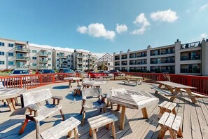 A wooden deck with picnic tables and benches, surrounded by a red railing, with modern buildings in the background. - Ski, Soak & Relax | Cozy Beech Mountain Condo (Beech Mountain)
