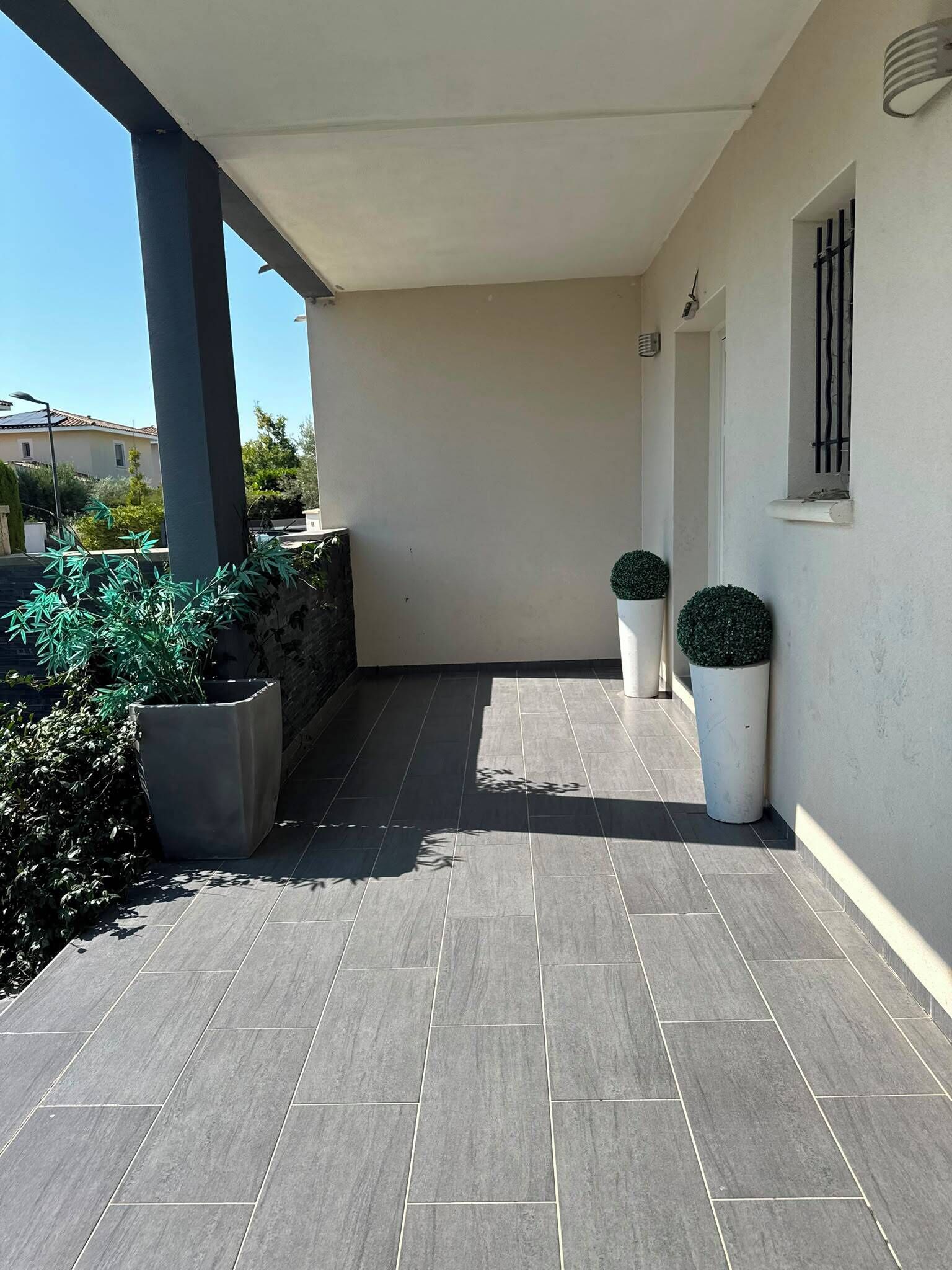 Zona exterior coberta amb paviment de rajoles, plantes en testos i entrada d'edifici.