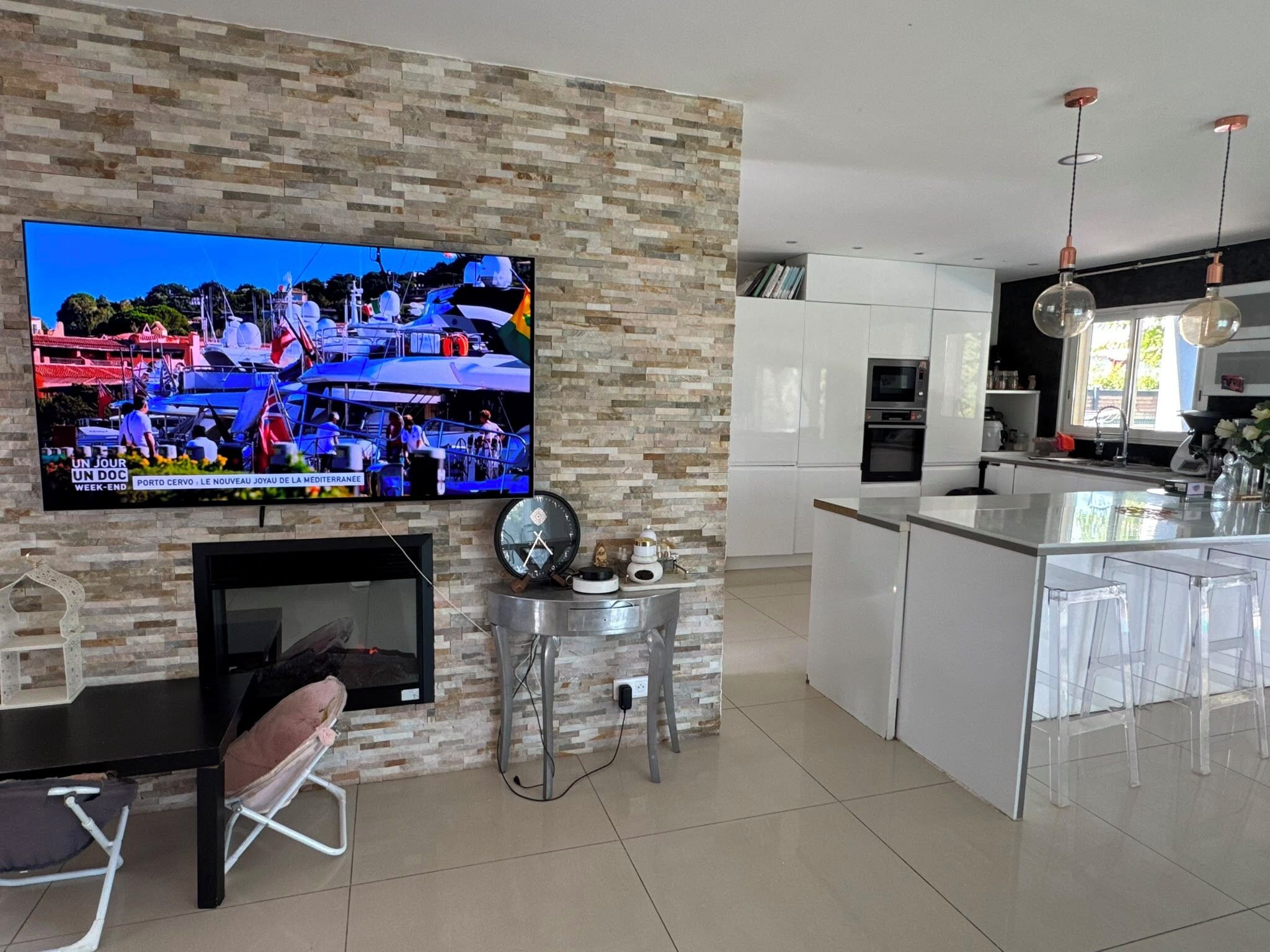 A modern kitchen with a TV mounted on a stone wall, a dining area with bar stools, and a small seating area with a chair and table.