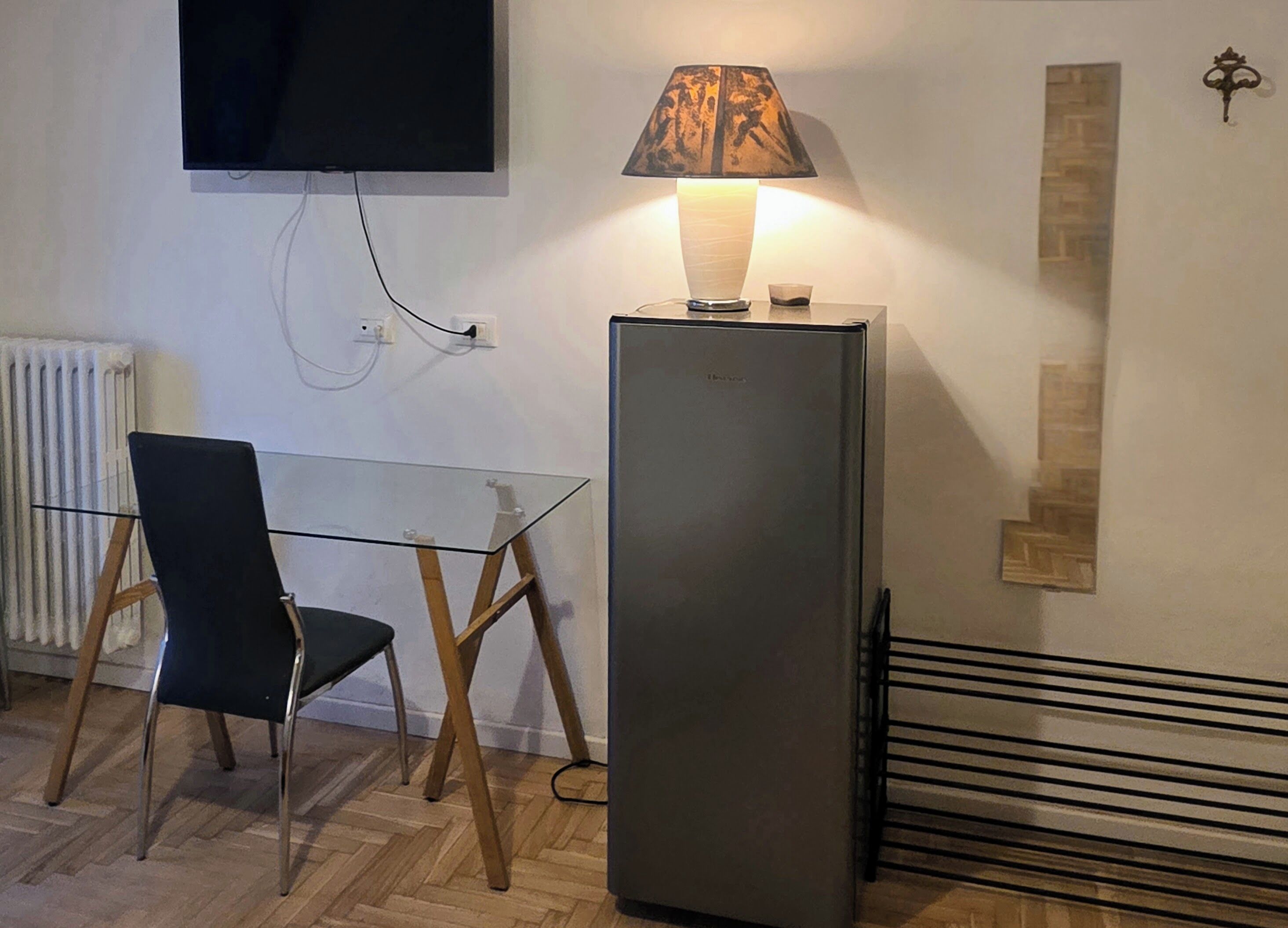 Una stanza con un tavolo in vetro, una sedia, una lampada da terra, una televisione a muro e un termosifone.