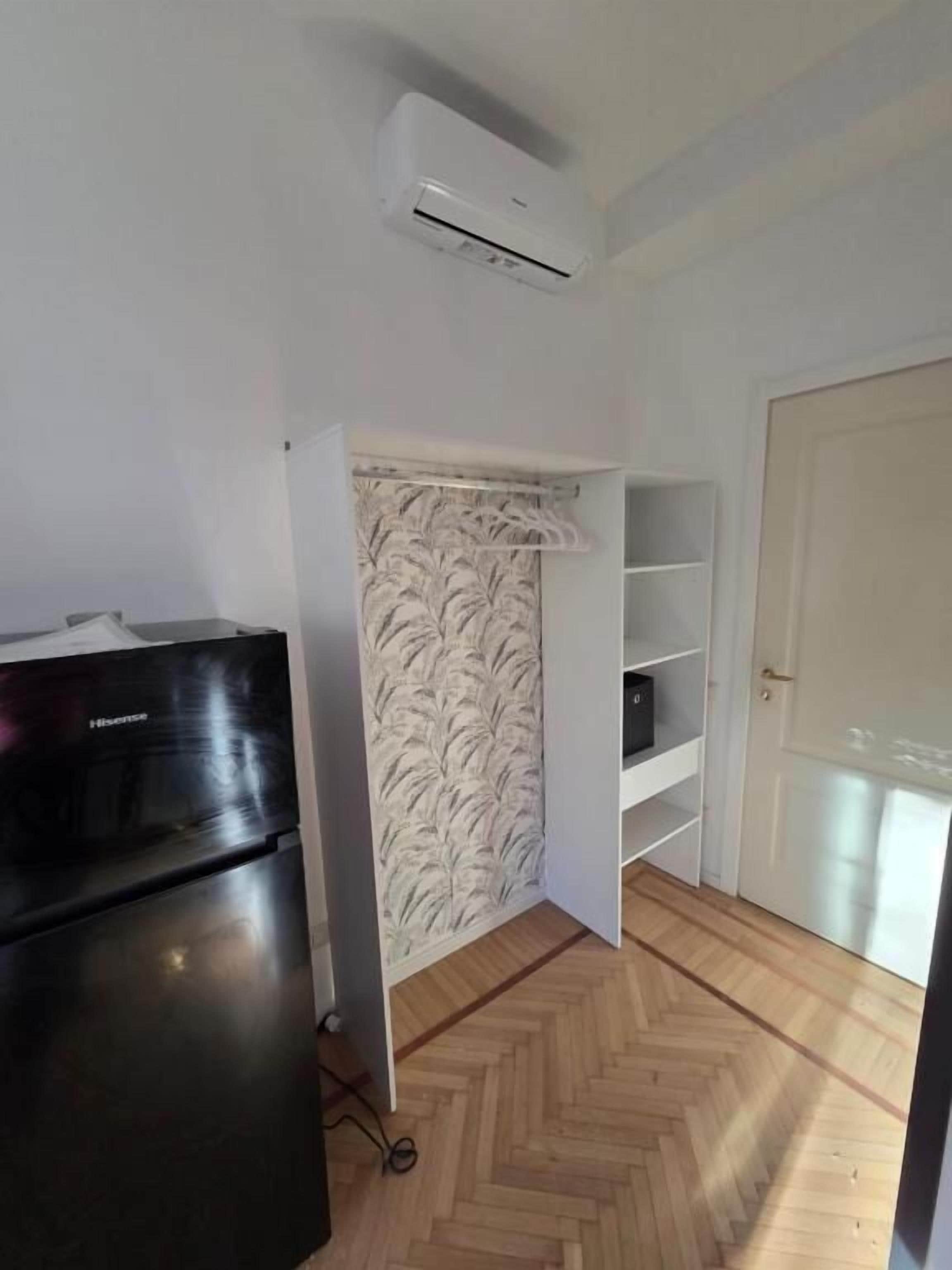 Ein Zimmer mit Kühlschrank, einer weißen Klimaanlage, einem gemusterten Vorhang und Holzboden.