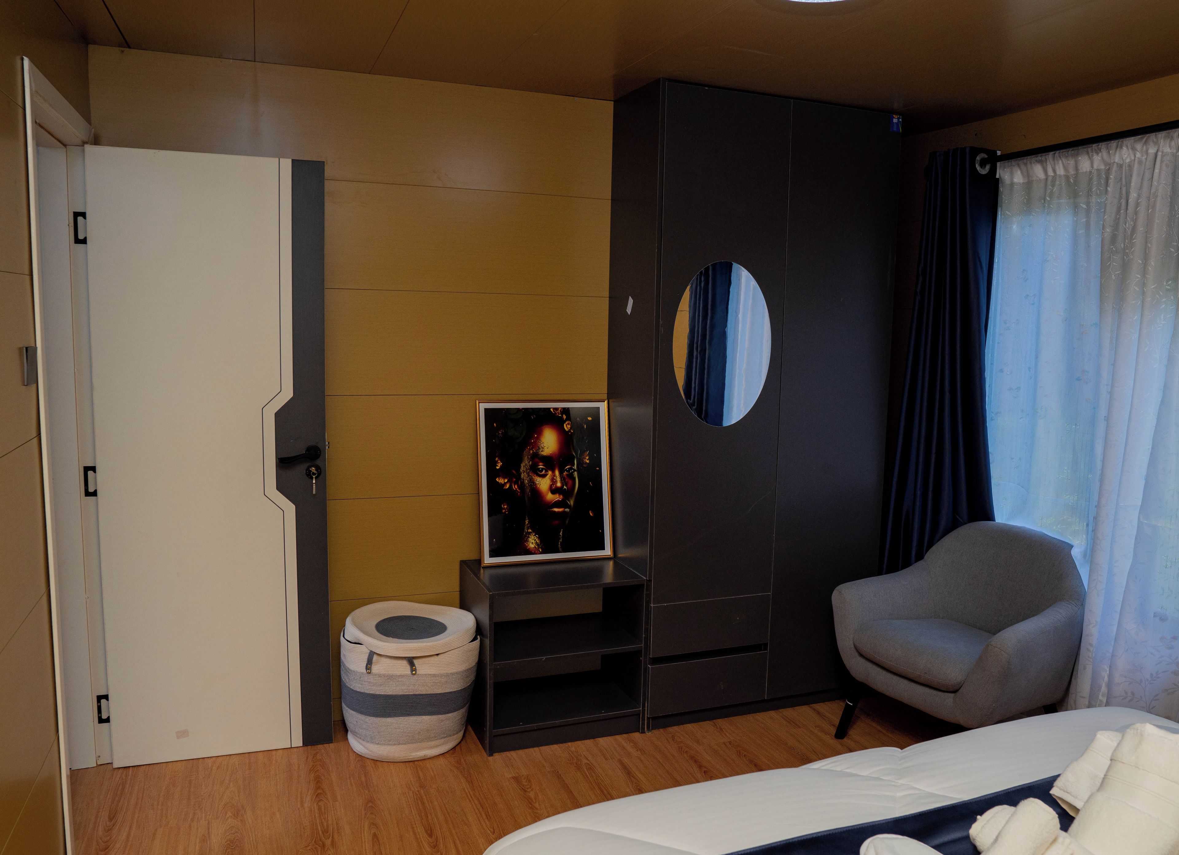 Ein Hotelzimmer mit einem Bett, einem grauen Sessel, einem kleinen Tisch mit einem gerahmten Bild, einem runden Spiegel und einem Kleiderschrank.