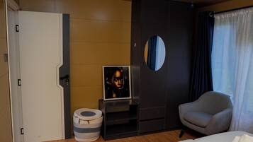A hotel room with a bed, a grey armchair, a small table with a framed picture, a round mirror, and a wardrobe.