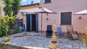 A stone patio with outdoor furniture, a potted plant, and a building with a pink facade. - Gisella's House Vacation Rental (Castellammare del Golfo)