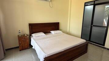 A hotel room with a wooden bed, a nightstand, an air conditioning unit, and a sliding door.