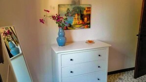 Een witte ladekast met drie lades, een blauwe vaas met roze bloemen en een schilderij aan de muur.