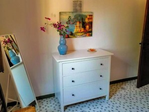 A white dresser with three drawers, a blue vase with pink flowers, and a painting on the wall. - Gisella's House (Sicilia)