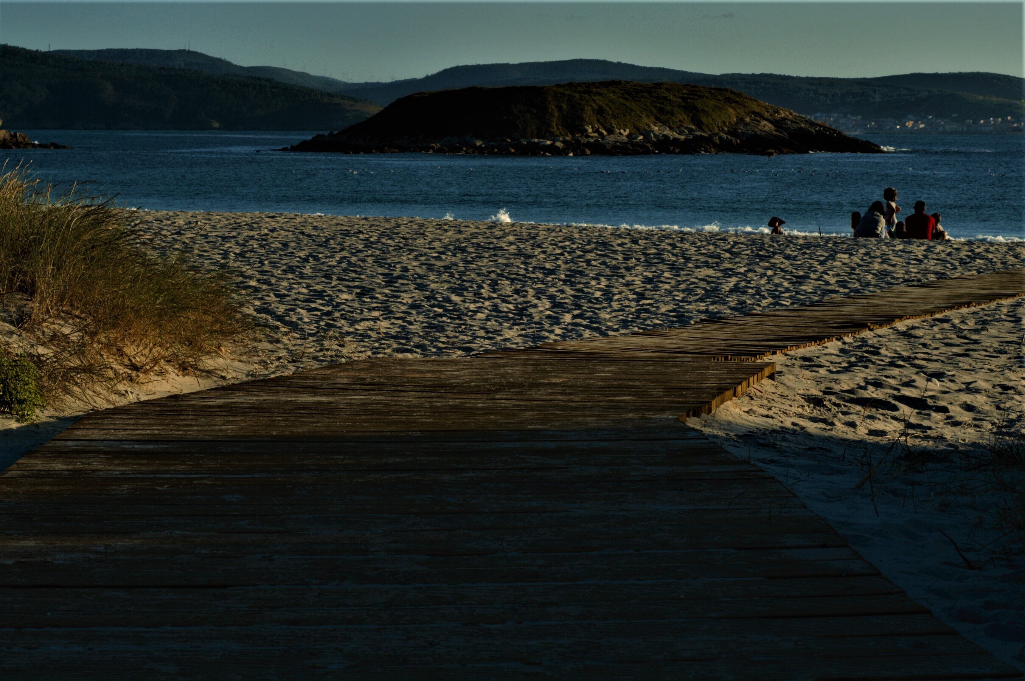 Un malecón de madera que conduce a una playa arenosa, con una pequeña isla en la distancia.