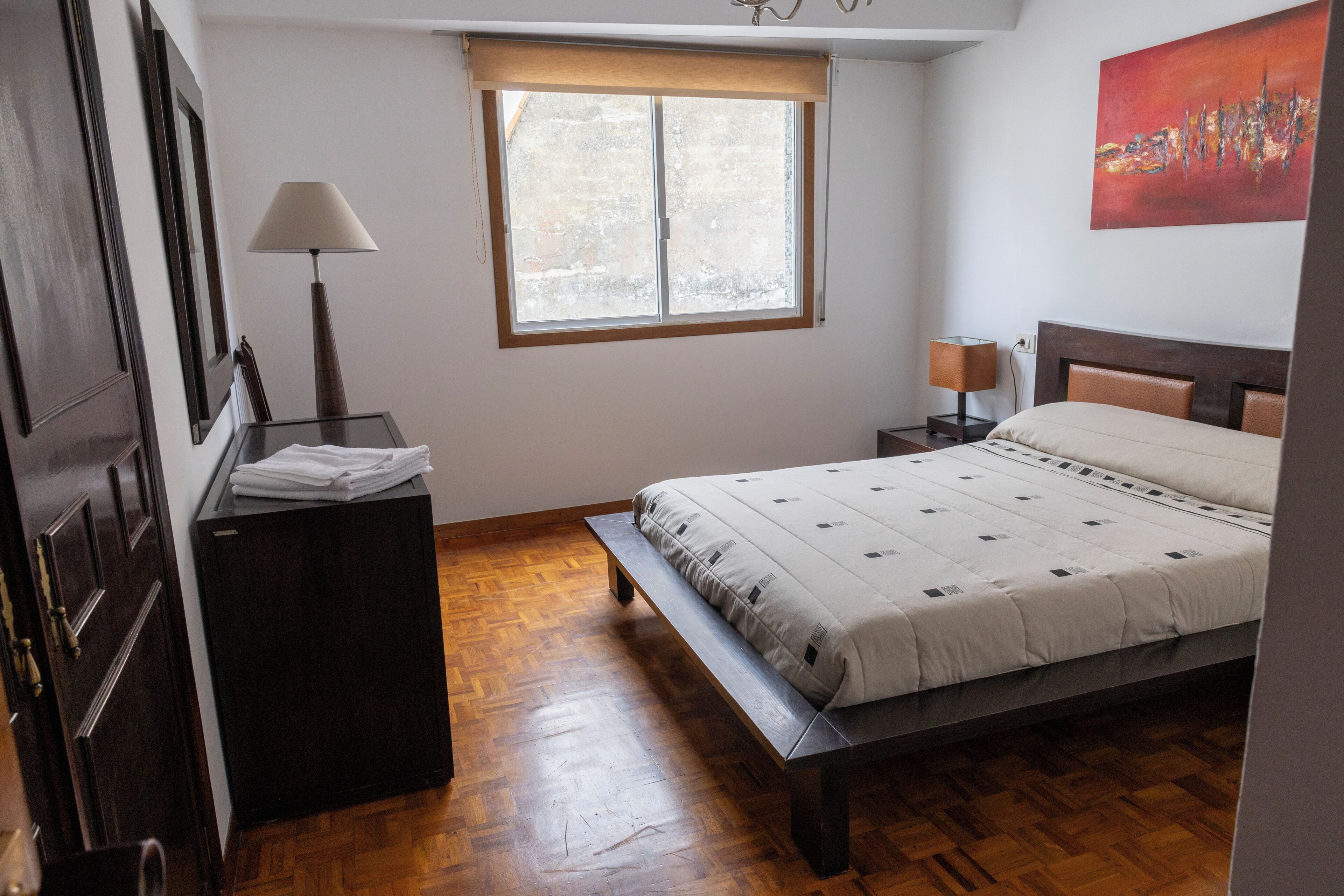 Un dormitorio con cama, escritorio, lámpara, ventana y un cuadro en la pared.