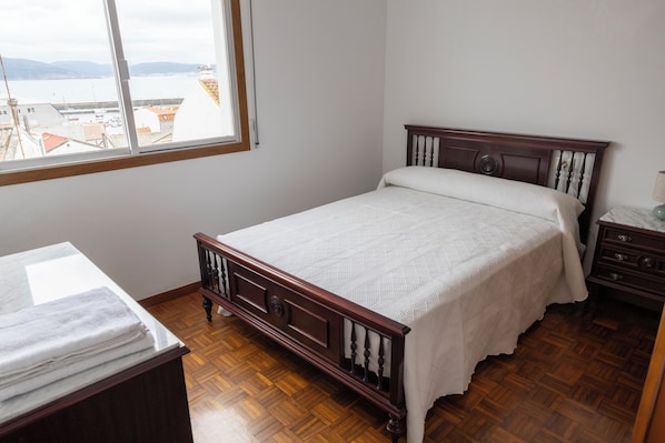 A bedroom with a wooden bed, a nightstand, a window with a view of buildings, and a wooden floor.