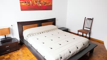 Un dormitorio ordenado con cama, mesita de noche, silla y un cuadro en la pared.